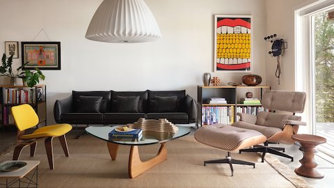A bright modern living room featuring colorful artwork on white walls and a white Nelson Bell Bubble Lamp over a lounge are with a yellow upholstered Eames LCW chair, a black leather Wireframe Sofa, and an Eames Lounge Chair and Ottoman upholstered in brown and white checkered Alexander Girard fabric around a glass and wood Noguchi Table. Eames Storage Units, an Eames Turned Stool, and Eames Hang-It-All complete the space.